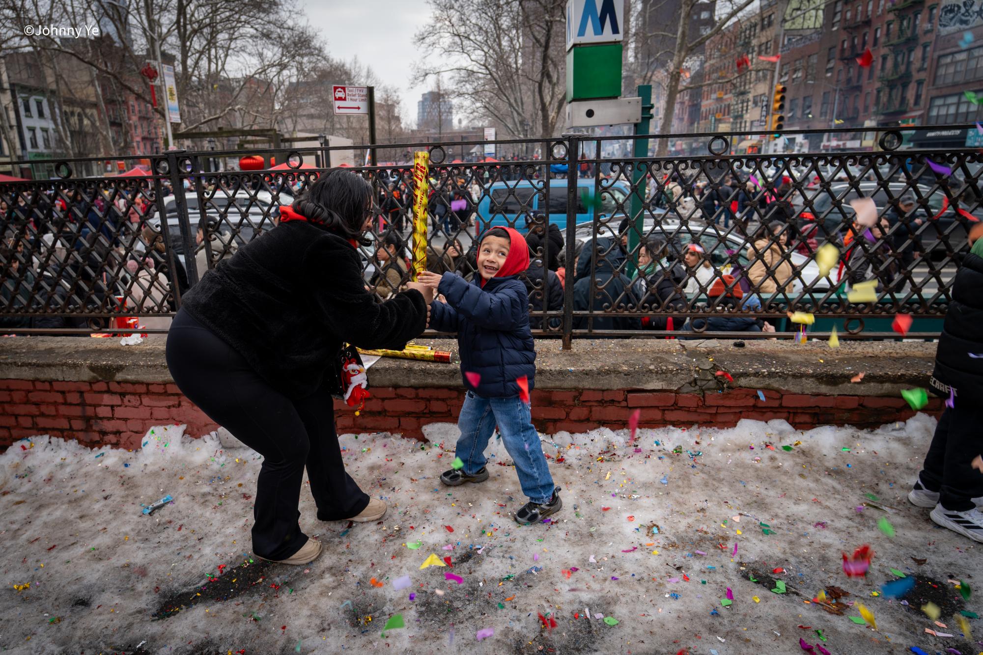 A boy smiles as he sets off the confetti popper, thanks to assistance from an adult, amidst celebrations of Lunar New Year in Chinatown, with crowds watching behind them, celebrating the arrival of the New Year. The celebration of the festival is marked by bringing people closer together through festivities such as parades and performances with symbolism of luck and prosperity. These bursts of colorful confetti resemble an ancient Lunar New Year tradition of driving out bad luck.
